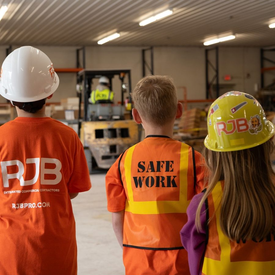 Kids in RJB shirts, hard hats, and safety vests watching a forklift in a warehouse on Bring Your Kid to Work Day